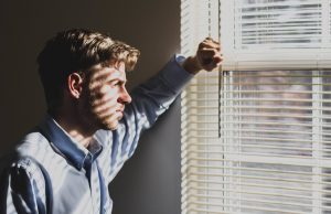 Image of a person leaning against the wall next to a window. The window's blinds are partway open and light is passing in through the window. The person looks pensive as they gaze out the window.