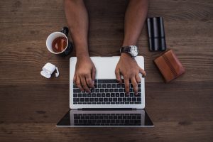 Image of a person's hands typing on a laptop. Next to the laptop, there is a cup of coffee, air pods, and a wallet.