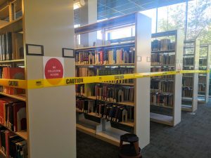 Image of a group of shelves that have yellow caution tape wrapped around them to prevent library users from browsing or touching the books. One of the shelves has a sign with the big red circle that reads "No Collection Access."