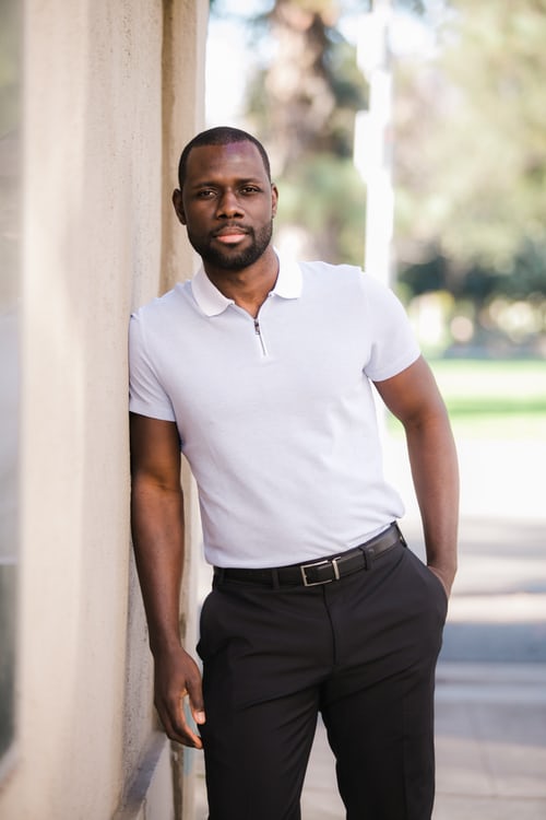 A man leans against a wall wearing a white polo with collar and slacks.