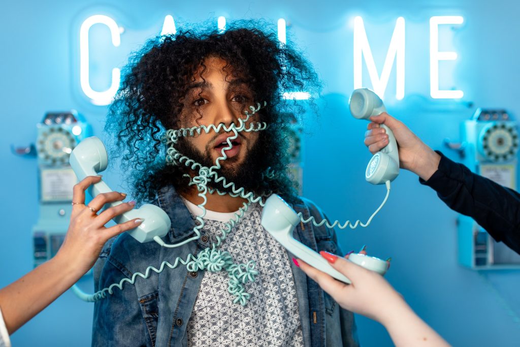 A man stands agape, entangled by phone cables. Behind him, a neon blue sign reads "CALL ME".