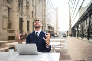 A man in a suit sits at a table with a laptop open. His face, upturned to the sky, shows unhappiness.