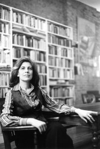 Photograph of Susan Sontag in 1979 seated on a wooden chair with bookcases behind her