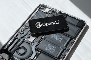 OpenAI logo displayed on a smartphone screen in landscape. The phone is laying on a laptop computer that has it's case open - revealing the internal batteries
