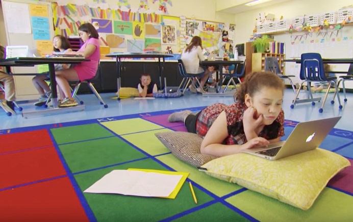 Image of a flexible learning space. Some students are at desks and one student is laying on the ground working on a laptop.