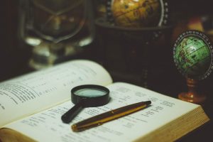 Open book with magnifier and pen laying on page and surrounded by tabletop globes.