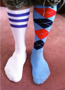 Two mismatched socks worn below the knee on a person. The right is a white sock with three blue bands along the top. The left sock is blue with a red, navy, and white argyle design on the leg portion.