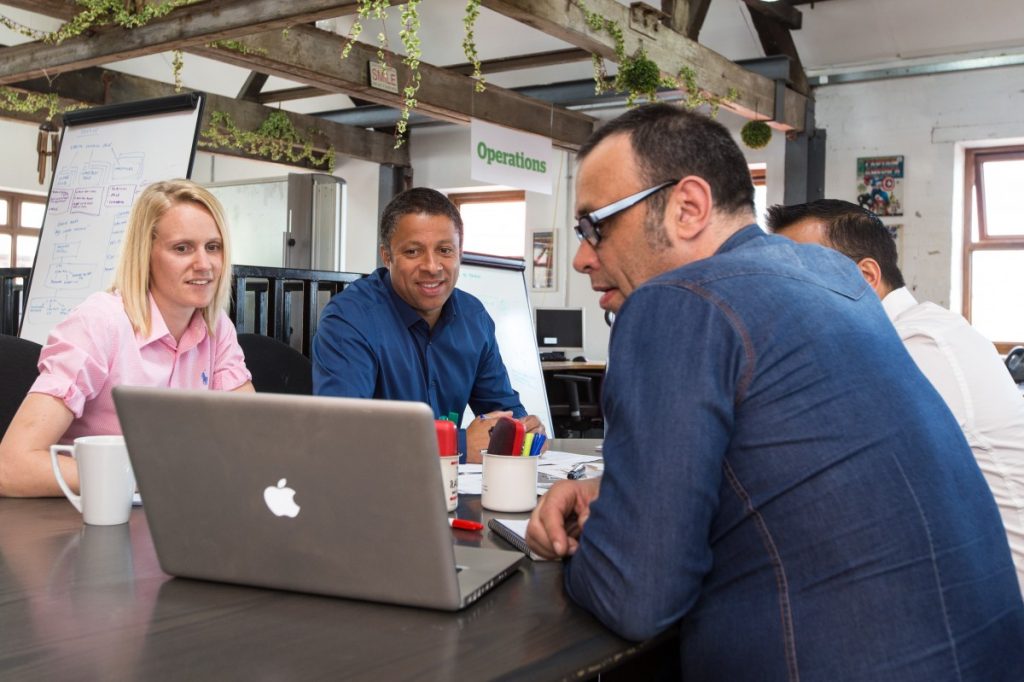 Descriptive image showing a group of young professionals, seated at a table and looking at a laptop.