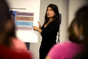 Descriptive image showing a woman speaking to an audience while pointing at a diagram.