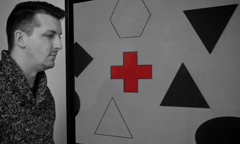 A photograph shows a person staring at a screen that displays one red cross toward the left side and numerous black and white shapes all over.