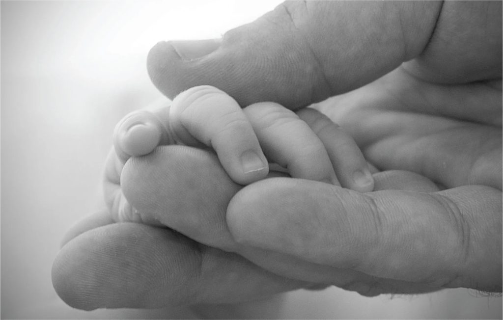 A picture shows two intertwined hands. One is the large hand of an adult, and the other is the tiny hand of an infant. The infant’s entire hand grasp is about the size of a single adult finger.